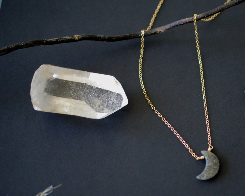 May include: A gold chain necklace with a crescent moon pendant. A clear quartz crystal and a dark twig are also in the image. The necklace is displayed on a dark background.