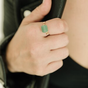 May include: A gold-colored ring featuring a rectangular, light green gemstone. The gemstone is the focal point, set in a delicate, wire-wrapped design. The ring is shown on a hand, with a black leather jacket in the background.