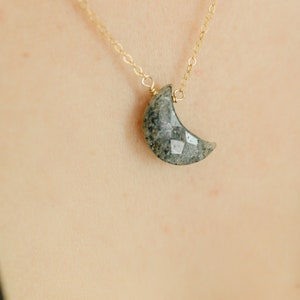 May include: A gold chain necklace with a crescent moon pendant. The moon pendant is a faceted, gray-green stone. The necklace is displayed against a light skin tone background.