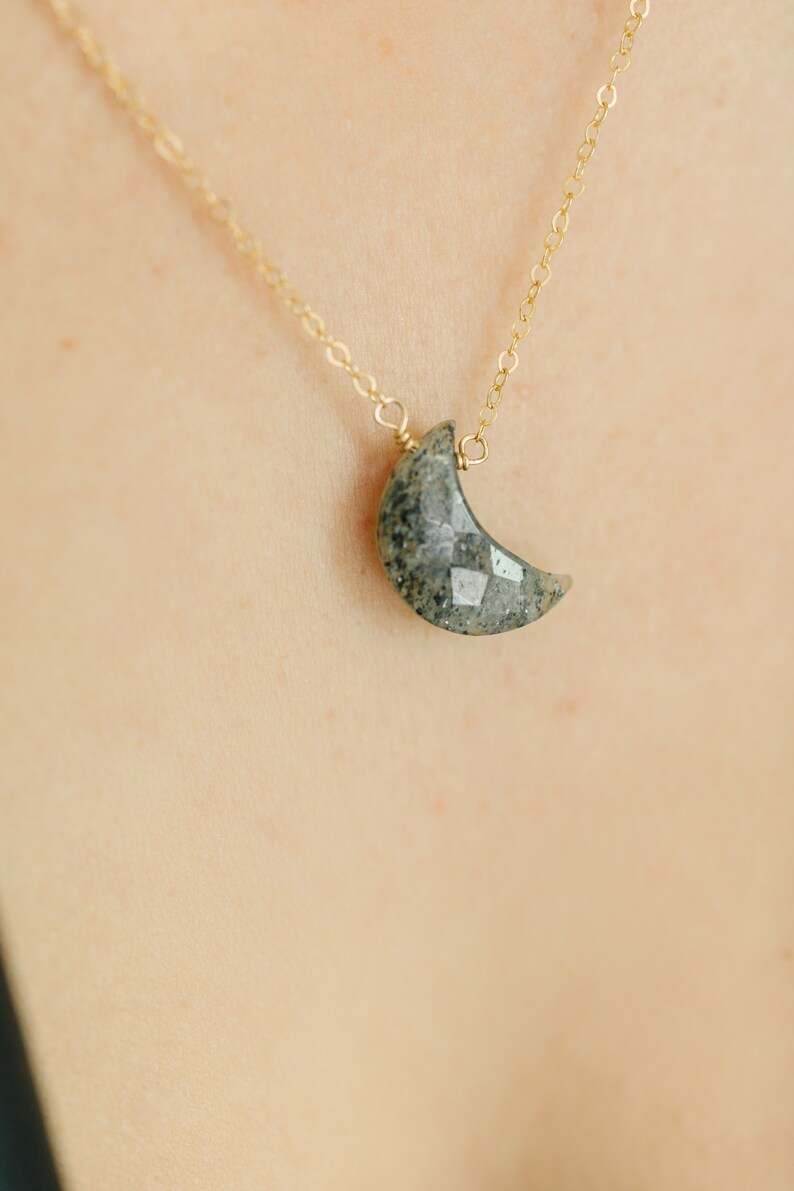 May include: A gold chain necklace with a crescent moon pendant. The moon pendant is a faceted, gray-green stone. The necklace is displayed against a light skin tone background.