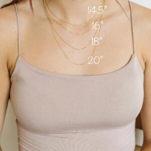 May include: A woman wearing a beige tank top with four gold necklaces of varying lengths: 14.5", 16", 18", and 20". The necklaces are displayed to show the different lengths. The image is well-lit and focuses on the necklaces.