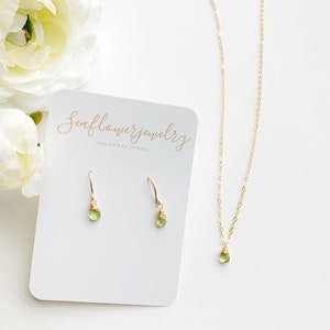 May include: A gold-colored necklace and earrings set with teardrop-shaped green gemstones. The earrings are hook-style, and the necklace has a delicate chain. A white card with the text "Seaflowerjewelry handmade jewelry" is also in the image.