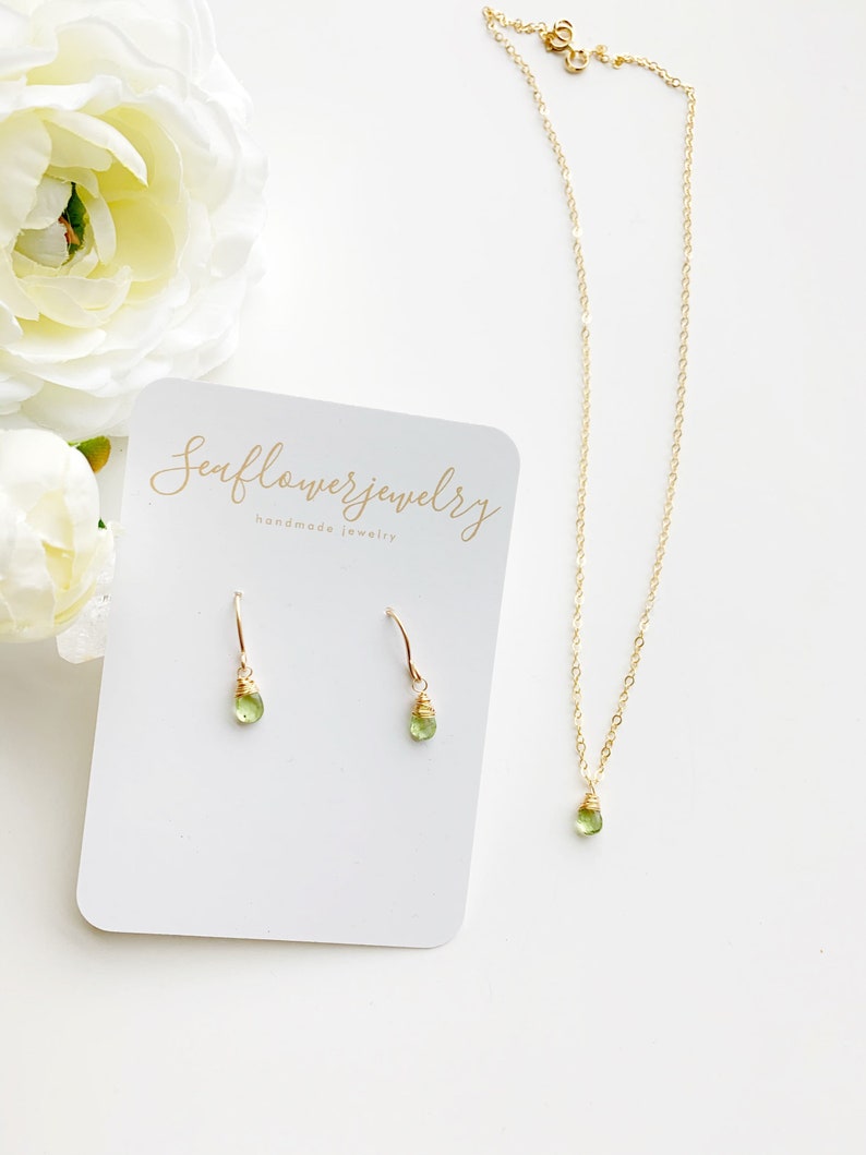 May include: A gold-colored necklace and earrings set with teardrop-shaped green gemstones. The earrings are hook-style, and the necklace has a delicate chain. A white card with the text "Seaflowerjewelry handmade jewelry" is also in the image.