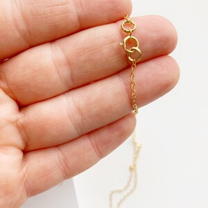 May include: Close-up of a delicate gold necklace with a small chain and clasp. The necklace is held in a hand, showcasing the fine details of the jewelry. The background is a soft, neutral color, highlighting the gold's shine.