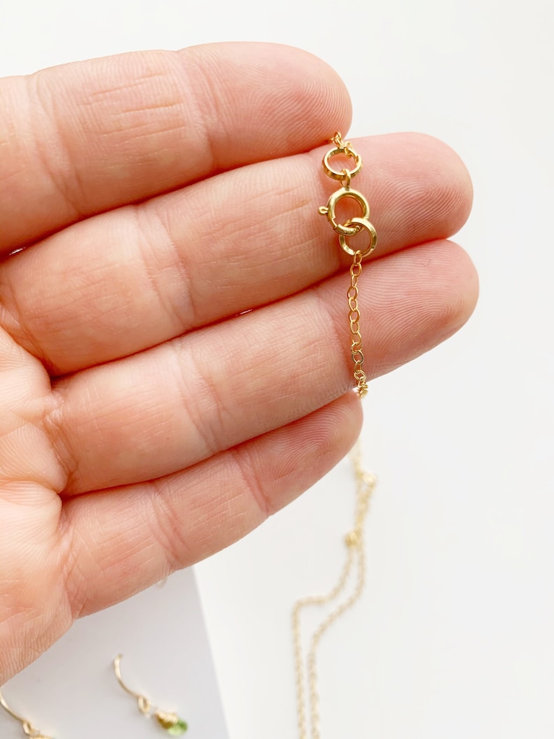 May include: Close-up of a delicate gold necklace with a small chain and clasp. The necklace is held in a hand, showcasing the fine details of the jewelry. The background is a soft, neutral color, highlighting the gold's shine.