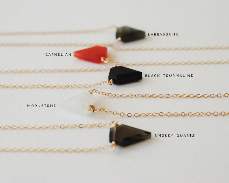 May include: Close-up of five delicate necklaces with gold chains and gemstone pendants. The gemstones include labradorite, carnelian, black tourmaline, moonstone, and smokey quartz. Each pendant is a different shape and color, with the names of the stones printed above.