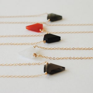 May include: A collection of delicate necklaces with gold chains. Each necklace features a geometric, triangular pendant in various colors: black, red, white, and dark green. The pendants are attached to the chain with a small gold loop.