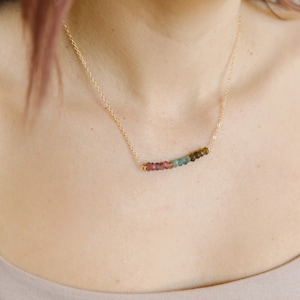May include: A delicate gold necklace featuring a row of colorful tourmaline beads. The beads transition in color from pink to green and brown. The necklace is worn on a person's neck, showcasing the jewelry's design.