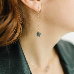 May include: A gold-colored earring with a star-shaped stone pendant. The earring hangs from a delicate chain. The image shows a close-up of the earring, highlighting its design and the texture of the stone. The earring is part of a jewelry set.