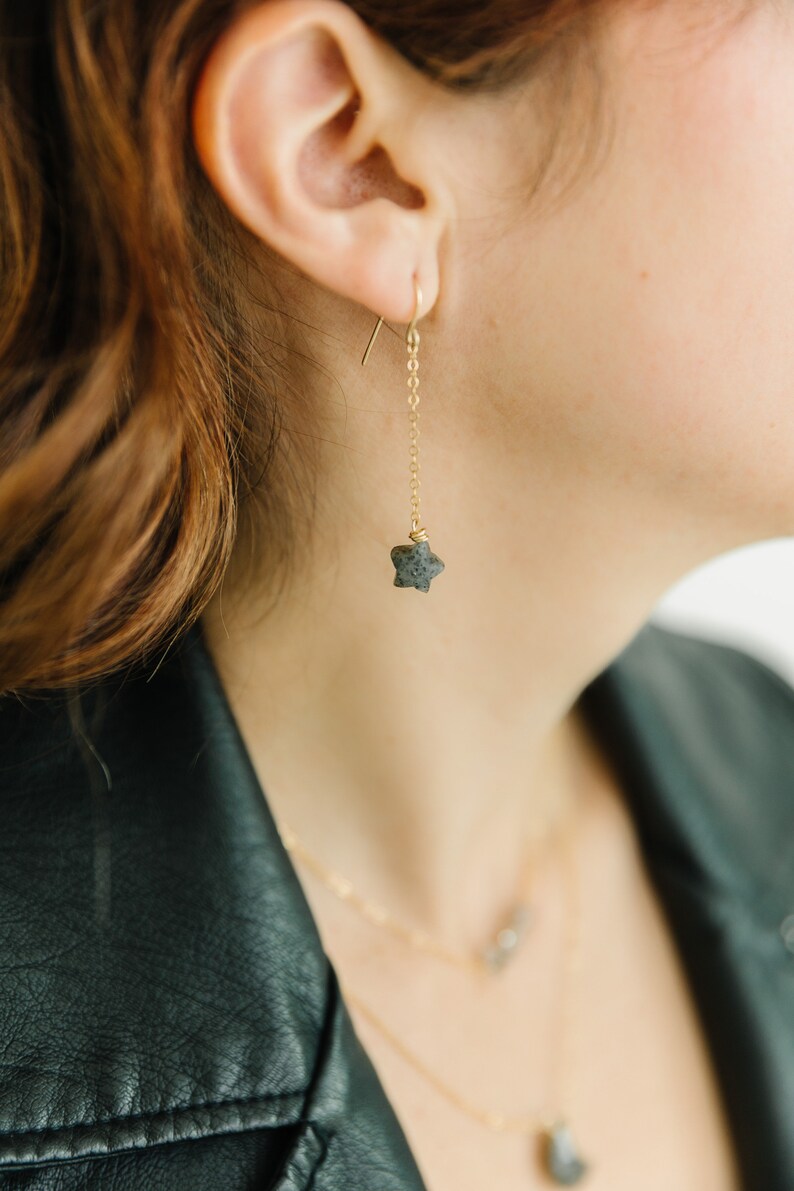 May include: A gold-colored earring with a star-shaped stone pendant. The earring hangs from a delicate chain. The image shows a close-up of the earring, highlighting its design and the texture of the stone. The earring is part of a jewelry set.