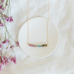May include: A delicate gold necklace featuring a row of colorful tourmaline beads. The beads transition from pink to green, creating a subtle gradient. The necklace rests on a white ceramic plate, with dried purple flowers in the background.