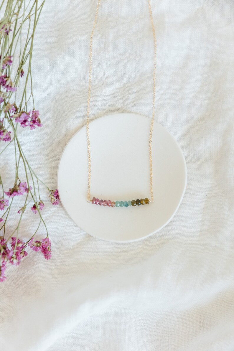 May include: A delicate gold necklace featuring a row of colorful tourmaline beads. The beads transition from pink to green, creating a subtle gradient. The necklace rests on a white ceramic plate, with dried purple flowers in the background.