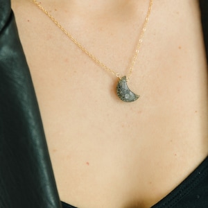 May include: A gold-colored chain necklace with a crescent moon pendant. The moon pendant is a dark gray color with a textured surface. The necklace is worn on a person wearing a black top and a black leather jacket.