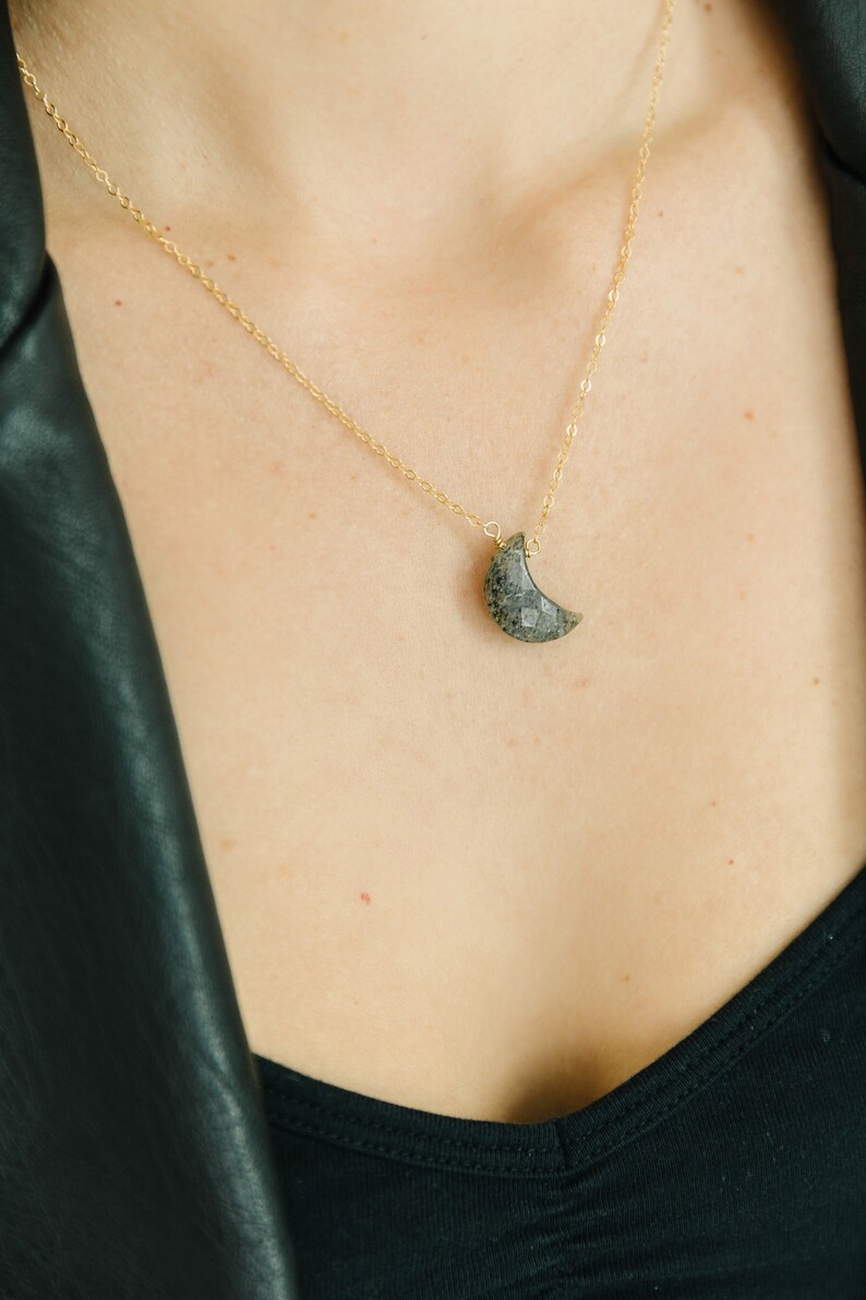 May include: A gold-colored chain necklace with a crescent moon pendant. The moon pendant is a dark gray color with a textured surface. The necklace is worn on a person wearing a black top and a black leather jacket.