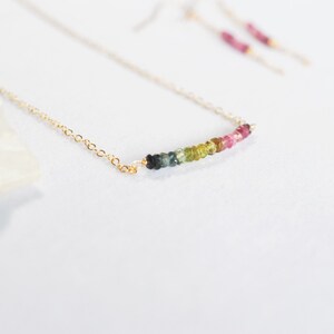 May include: A delicate gold necklace with a bar of colorful tourmaline beads. The necklace features a thin gold chain and a row of green, yellow, pink, and purple gemstones. Matching earrings are visible in the background.