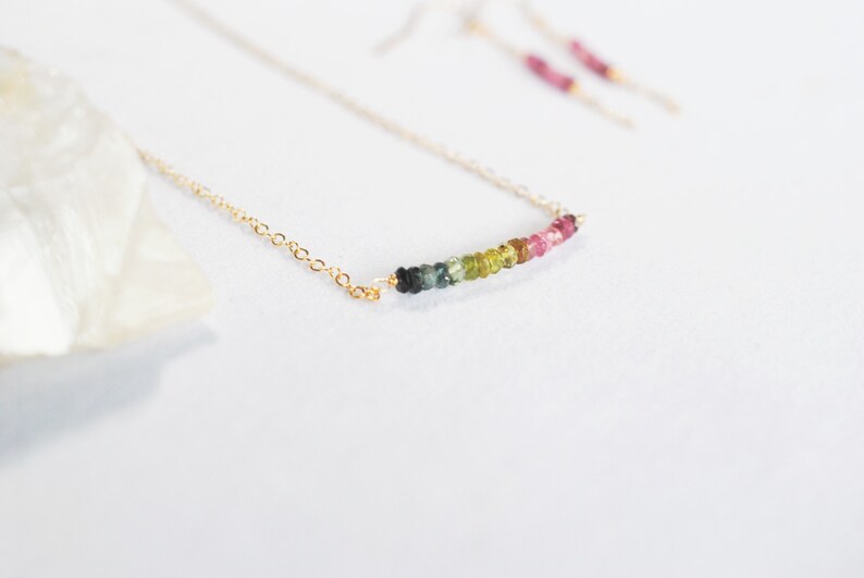 May include: A delicate gold necklace with a bar of colorful tourmaline beads. The necklace features a thin gold chain and a row of green, yellow, pink, and purple gemstones. Matching earrings are visible in the background.