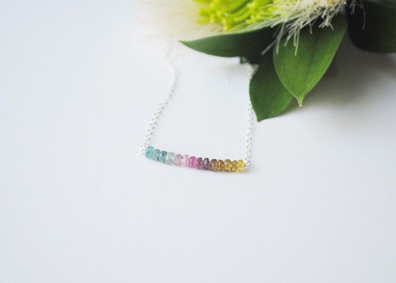 May include: A delicate necklace featuring a strand of colorful tourmaline beads in shades of blue, pink, and yellow. The gemstones are strung on a fine silver chain. The necklace is displayed on a white surface with green foliage in the background.