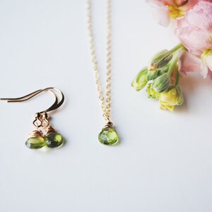 May include: A gold-toned jewelry set featuring a pair of drop earrings and a necklace with a single pendant. Each piece showcases a pear-shaped, light green gemstone. Soft pink and purple flowers are in the background.