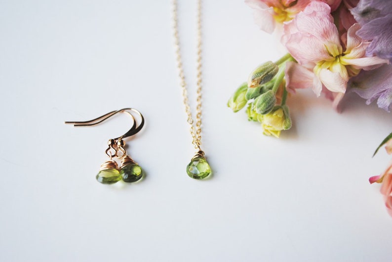 May include: A gold-toned jewelry set featuring a pair of drop earrings and a necklace with a single pendant. Each piece showcases a pear-shaped, light green gemstone. Soft pink and purple flowers are in the background.
