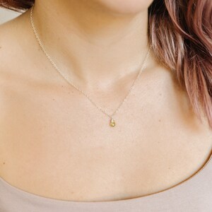May include: A delicate silver necklace with a small, pear-shaped, light green gemstone pendant. The necklace is worn on a person's neck, with the pendant resting near the collarbone. The background is a neutral color.