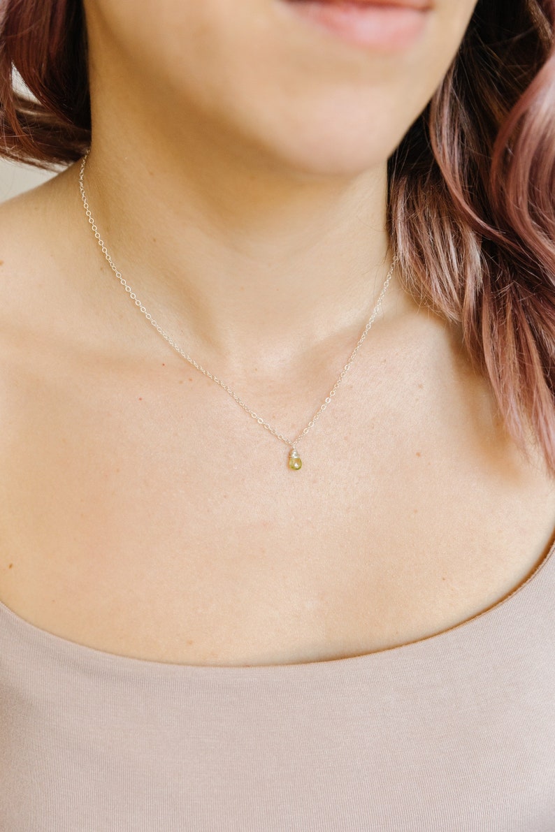 May include: A delicate silver necklace with a small, pear-shaped, light green gemstone pendant. The necklace is worn on a person's neck, with the pendant resting near the collarbone. The background is a neutral color.