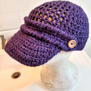 May include: A purple crocheted hat with a visor and a wooden button. The hat has an open weave design on the top and a solid crochet brim. The hat is displayed on a white mannequin head.