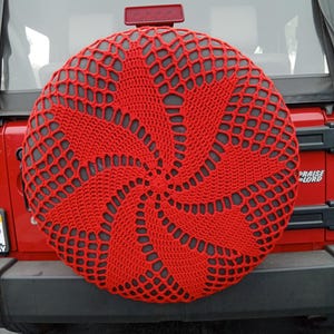 Pode incluir: Capa vermelha de crochê para pneu sobressalente com design em espiral. A capa, feita de material semelhante a uma rede, é montada na parte traseira de um veículo vermelho. O design é centralizado e cobre todo o pneu.