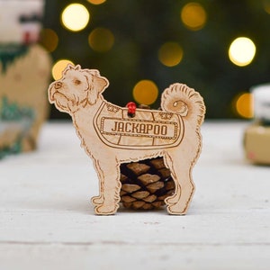 May include: A wooden Christmas bauble shaped like a dog with the name "Jackapoo" engraved on it. The bauble is painted in a light wood colour and has a red string attached to the top.