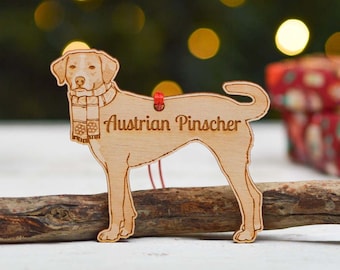 May include: Wooden ornament in the shape of an Austrian Pinscher dog, wearing a scarf. The dog is light brown with the text "Austrian Pinscher" engraved on the body. A red string is attached for hanging. The ornament is resting on a wooden branch.