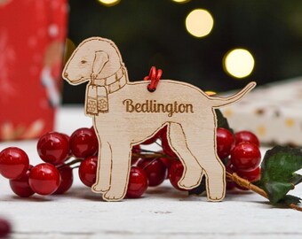 May include: A wooden Christmas ornament shaped like a Bedlington Terrier dog wearing a scarf. The ornament is light brown and has the word "Bedlington" engraved on it.