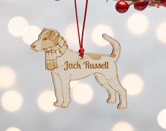 May include: Wooden Christmas ornament shaped like a Jack Russell terrier wearing a scarf. The ornament is hanging from a red string and has the words "Jack Russell" engraved on it.