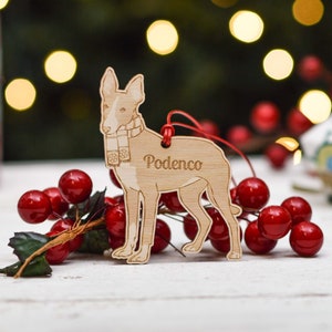 May include: A wooden Christmas tree decoration shaped like a dog wearing a scarf. The decoration is engraved with the name "Podenco".