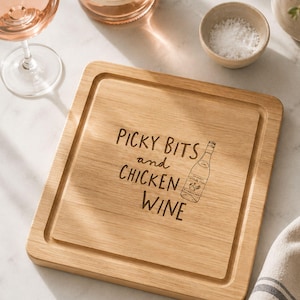May include: A square wooden serving board with a grooved border. The centre is laser-engraved with the text "PICKY BITS and CHICKEN WINE" alongside a line-art illustration of a wine bottle. The board features a natural light brown wood grain.
