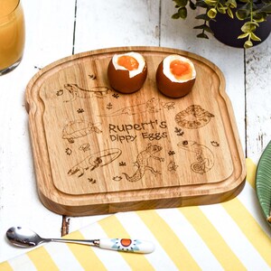 May include: A wooden breadboard shaped like a slice of bread with a personalised inscription "Rupert's Dippy Eggs" and a design of lizards, turtles, and leaves. Two soft-boiled eggs are on the board.