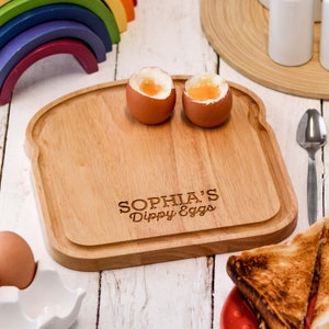 May include: A wooden bread-shaped serving tray with the text "Sophia's Dippy Eggs" engraved on it. Two soft-boiled eggs are on the tray, and a piece of toast is on a red plate in the background.