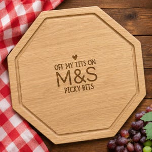 May include: An octagonal wooden serving board with a grooved border. The centre is laser-engraved with a small heart icon above the text "OFF MY TITS ON M&S PICKY BITS" in a clean, sans-serif font. The board features a natural light wood grain.