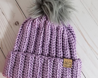 Peut inclure: Un bonnet en crochet violet avec un pompon en fausse fourrure gris et une étiquette en cuir marron avec les lettres "MC" dessus.