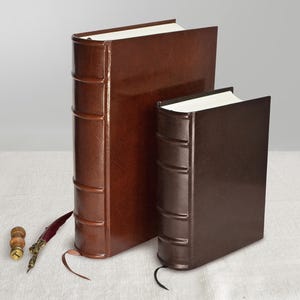 Puede incluir: Dos libros de cuero marrón de diferentes tamaños. El libro más grande tiene un diseño vertical con bandas en relieve. Una pluma y un pequeño sello de madera con detalles dorados están colocados cerca de los libros.