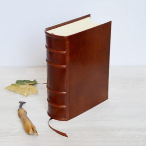 May include: A brown leather-bound book with a raised spine featuring five horizontal bands, cream-colored pages, and a thin brown ribbon bookmark extending from the bottom. A small wooden tool and a single dried leaf are placed beside the book.