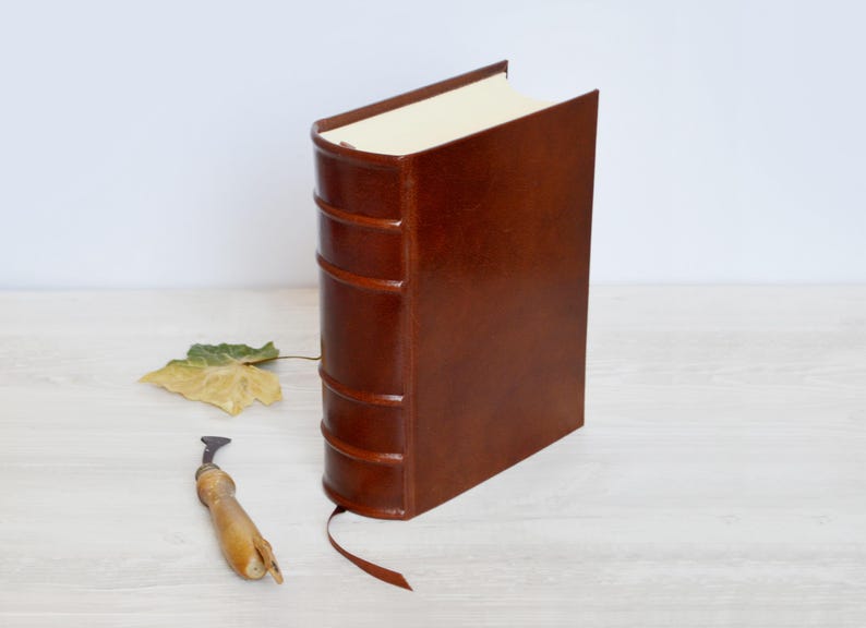 May include: A brown leather-bound book with a raised spine featuring five horizontal bands, cream-colored pages, and a thin brown ribbon bookmark extending from the bottom. A small wooden tool and a single dried leaf are placed beside the book.