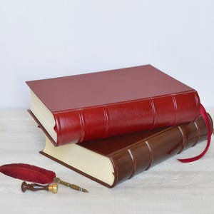 Peut inclure: Deux livres à couverture rigide empilés, l'un rouge foncé et l'autre marron foncé, tous deux dotés de nerfs sur le dos et de pages couleur crème. Une plume d'oie rouge avec une pointe en métal doré et un sceau à cire sont posés à côté.