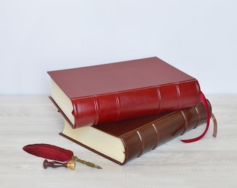 May include: Two hardcover books stacked on top of each other, one in deep red and one in dark brown, both featuring raised spine bands and cream-colored pages. A red feather quill pen with a brass-colored metal tip and a wax seal stamp are placed nearby.