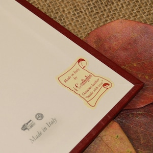 May include: A white label with red text that reads "Made in Italy by Il Carliglio Genuine leather "made with love"". The label is attached to a red leather notebook.