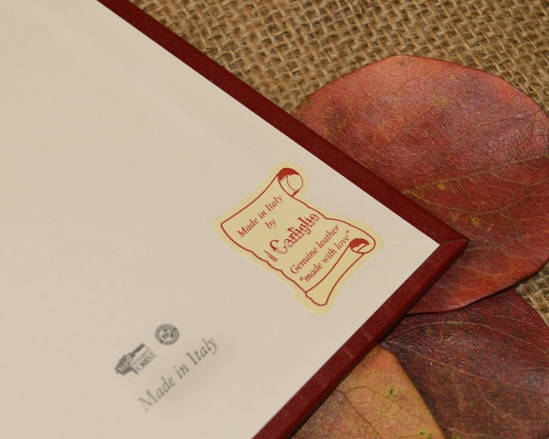 May include: A white label with red text that reads "Made in Italy by Il Carliglio Genuine leather "made with love"". The label is attached to a red leather notebook.