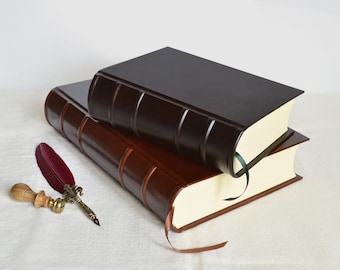 May include: Two leather-bound books, one dark brown and one lighter brown, stacked. A burgundy feather quill and a wooden stamp with a brass accent are next to the books. The books have cream-coloured pages.