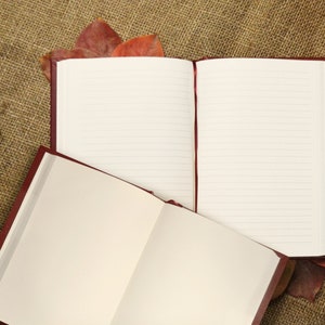May include: Two open burgundy leather journals with lined pages, lying on a brown burlap surface with dried leaves.