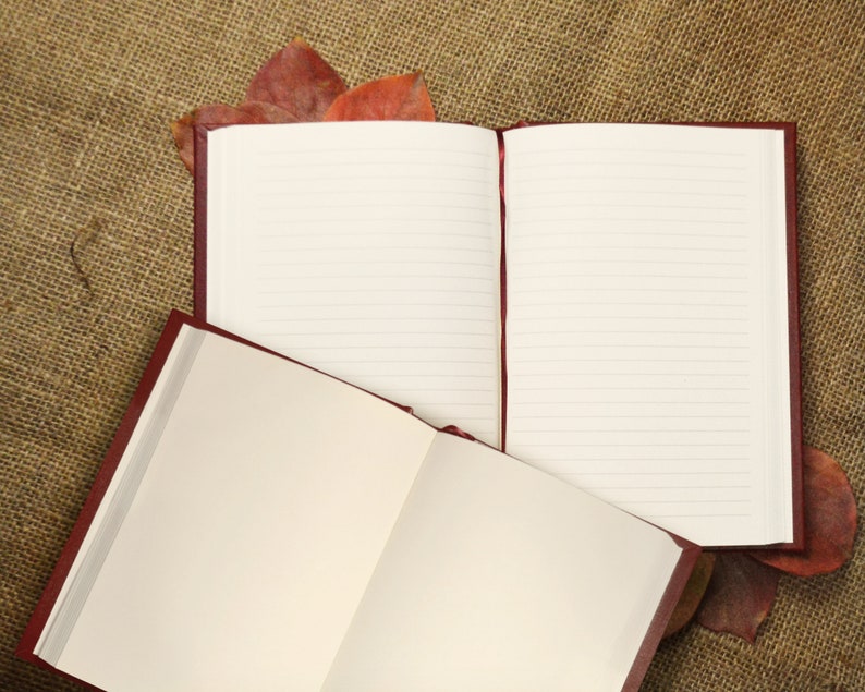 May include: Two open burgundy leather journals with lined pages, lying on a brown burlap surface with dried leaves.
