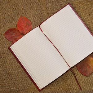 May include: A red leather-bound journal with lined pages is open and lying on a brown burlap surface.  Dried red leaves are scattered around the journal.