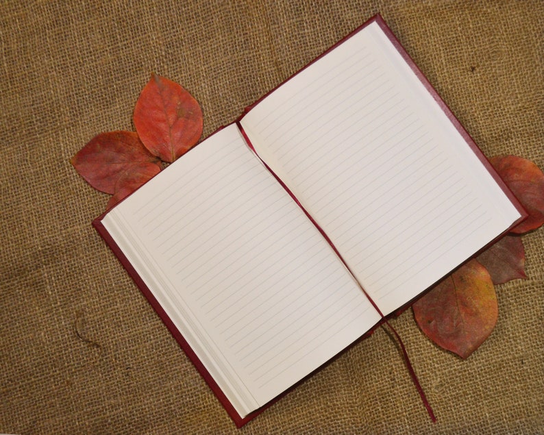 May include: A red leather-bound journal with lined pages is open and lying on a brown burlap surface.  Dried red leaves are scattered around the journal.
