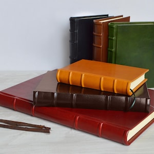 May include: A stack of five leather-bound journals in various colors, including red, brown, orange, green, and black. The journals have decorative raised lines and are arranged in a pyramid shape.